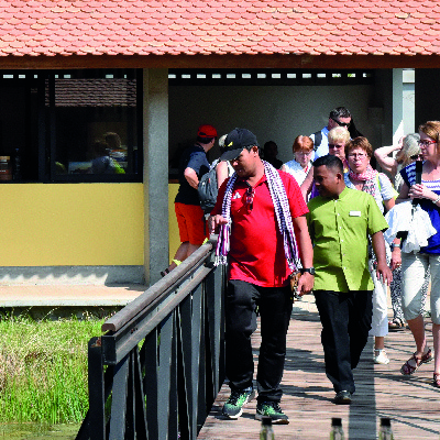 Visitors taking a guided tour at Senteurs d'Angkor, crossing a wooden bridge with a guide in a green shirt. They are exploring the workshop area surrounded by lush greenery.