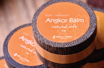 Stacked containers of Angkor Balm with orange labels that read 'pain reliever, natural soft, 20g' in white text, displayed on a wooden surface. The balm is marketed as a natural pain relief product.